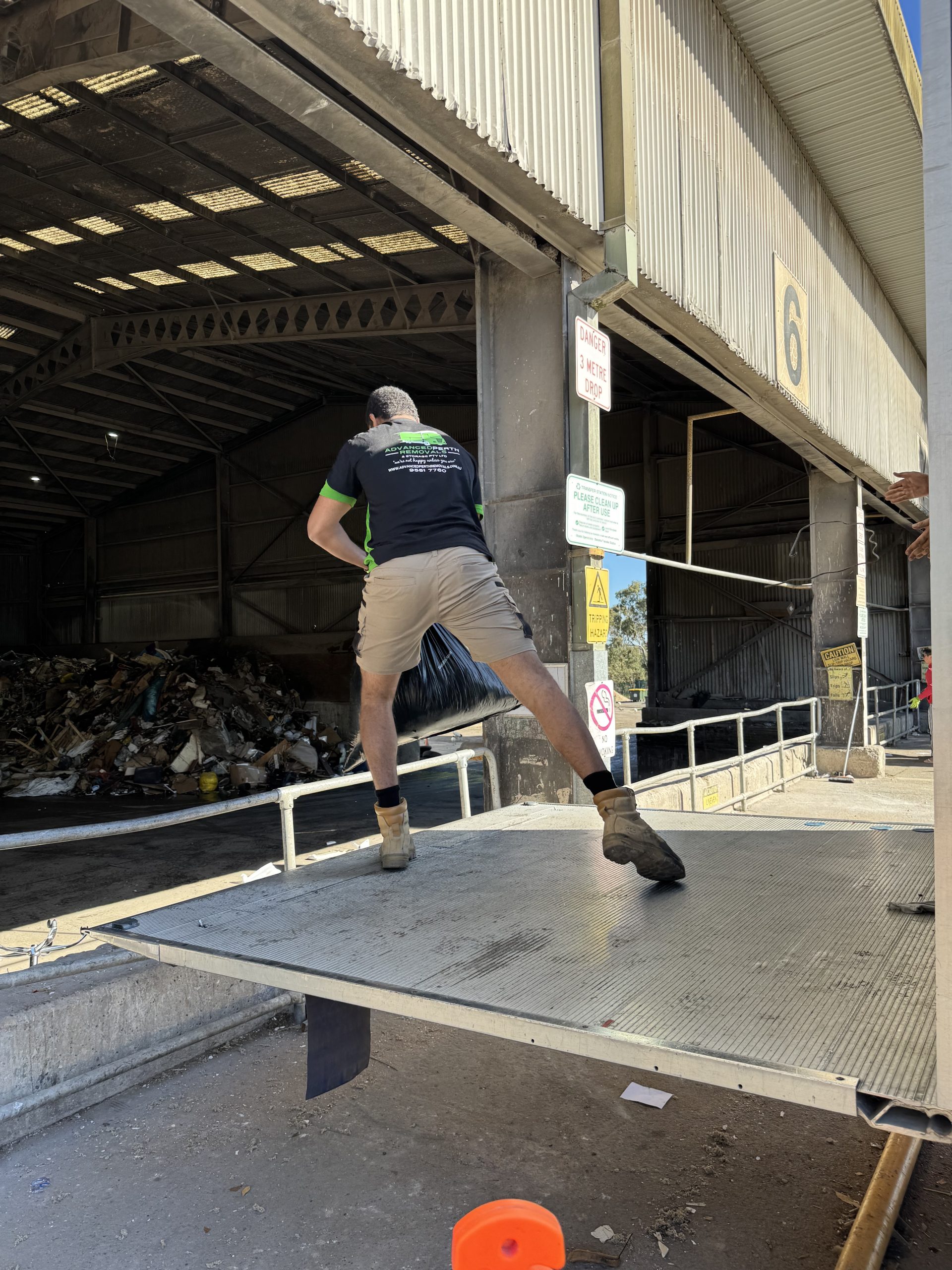 Man wearing work clothes leaps on a loading platform while carrying a large black garbage bag near an industrial building.